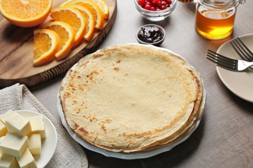 Clatite clasice asezate pe farfurie, alaturi de unt, fructe si dulceata